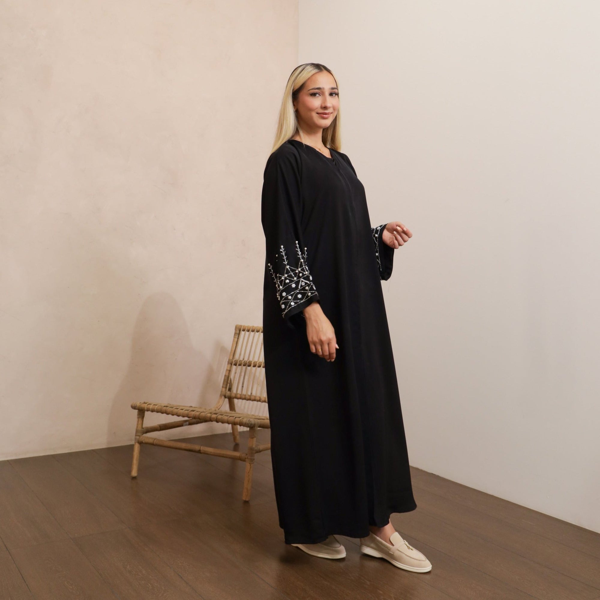 Black abaya in sizes 50-60 with hand-sewn silver and white pearl-like beadwork on the arms. Made from Zoom fabric, it features popper buttons and includes a matching black hijab. Handwash to preserve beadwork.