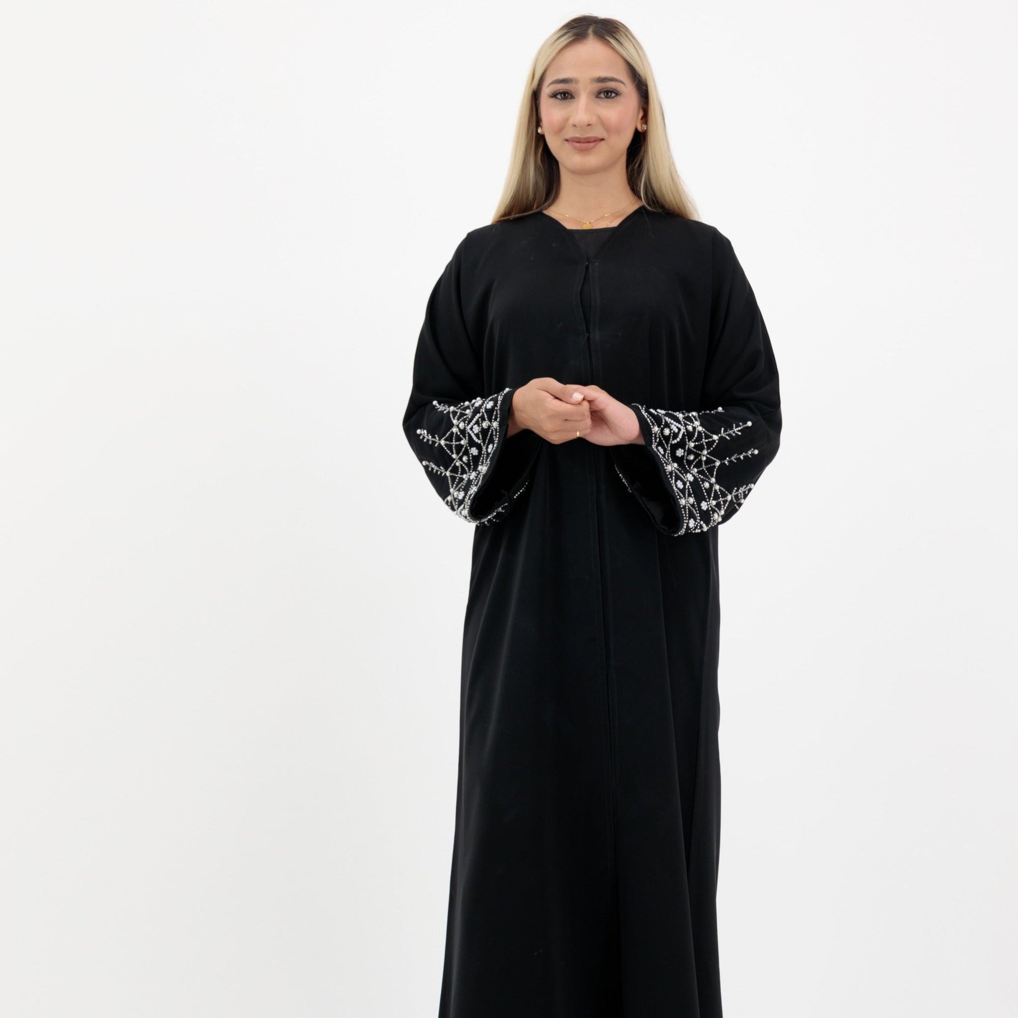Black abaya in sizes 50-60 with hand-sewn silver and white pearl-like beadwork on the arms. Made from Zoom fabric, it features popper buttons and includes a matching black hijab. Handwash to preserve beadwork.