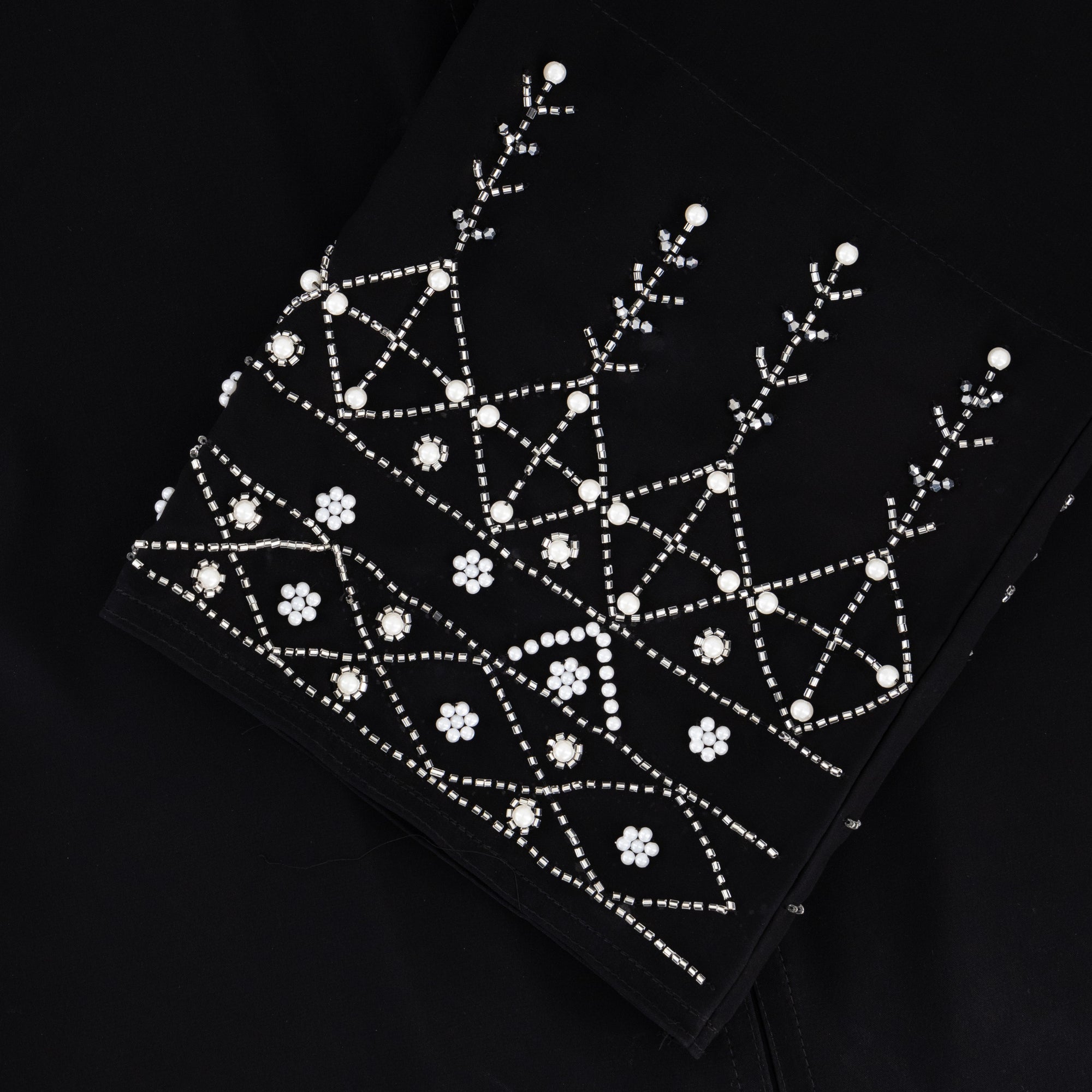 Black abaya in sizes 50-60 with hand-sewn silver and white pearl-like beadwork on the arms. Made from Zoom fabric, it features popper buttons and includes a matching black hijab. Handwash to preserve beadwork.