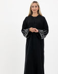 Black abaya in sizes 50-60 with hand-sewn silver and white pearl-like beadwork on the arms. Made from Zoom fabric, it features popper buttons and includes a matching black hijab. Handwash to preserve beadwork.