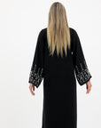Black abaya in sizes 50-60 with hand-sewn silver and white pearl-like beadwork on the arms. Made from Zoom fabric, it features popper buttons and includes a matching black hijab. Handwash to preserve beadwork.