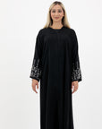 Black abaya in sizes 50-60 with hand-sewn silver and white pearl-like beadwork on the arms. Made from Zoom fabric, it features popper buttons and includes a matching black hijab. Handwash to preserve beadwork.
