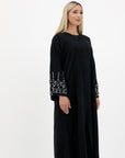 Black abaya in sizes 50-60 with hand-sewn silver and white pearl-like beadwork on the arms. Made from Zoom fabric, it features popper buttons and includes a matching black hijab. Handwash to preserve beadwork.