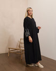 Black abaya in sizes 50-60 with hand-sewn silver and white pearl-like beadwork on the arms. Made from Zoom fabric, it features popper buttons and includes a matching black hijab. Handwash to preserve beadwork.