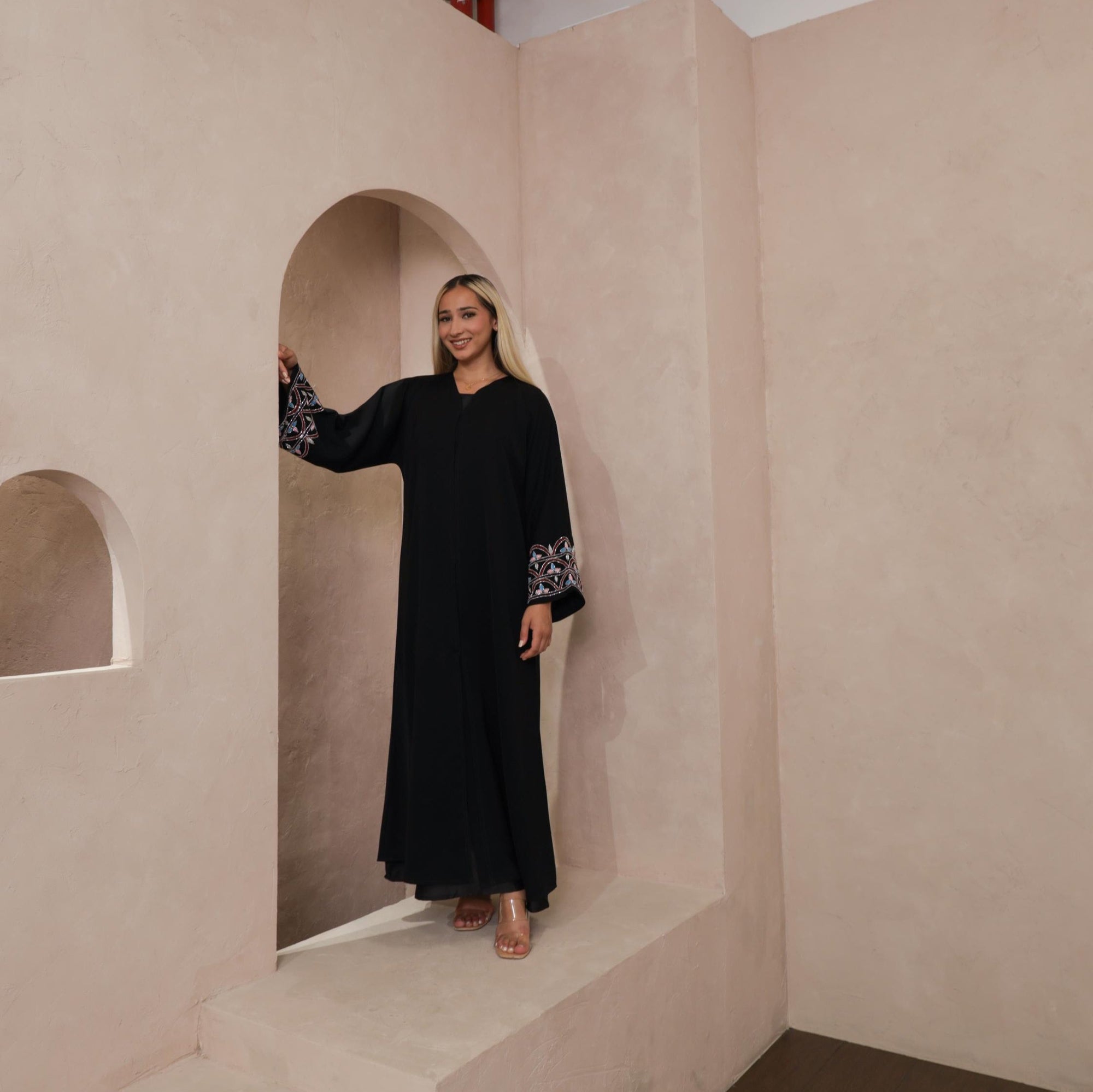 Black Liana Abaya with hand-sewn silver beads in a leaf and embroidery design pattern on the arms. Light pink, blue, and purple threads add color to the embroidery. Comes with a matching black hijab and popper buttons for closure.
