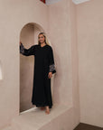Black Liana Abaya with hand-sewn silver beads in a leaf and embroidery design pattern on the arms. Light pink, blue, and purple threads add color to the embroidery. Comes with a matching black hijab and popper buttons for closure.