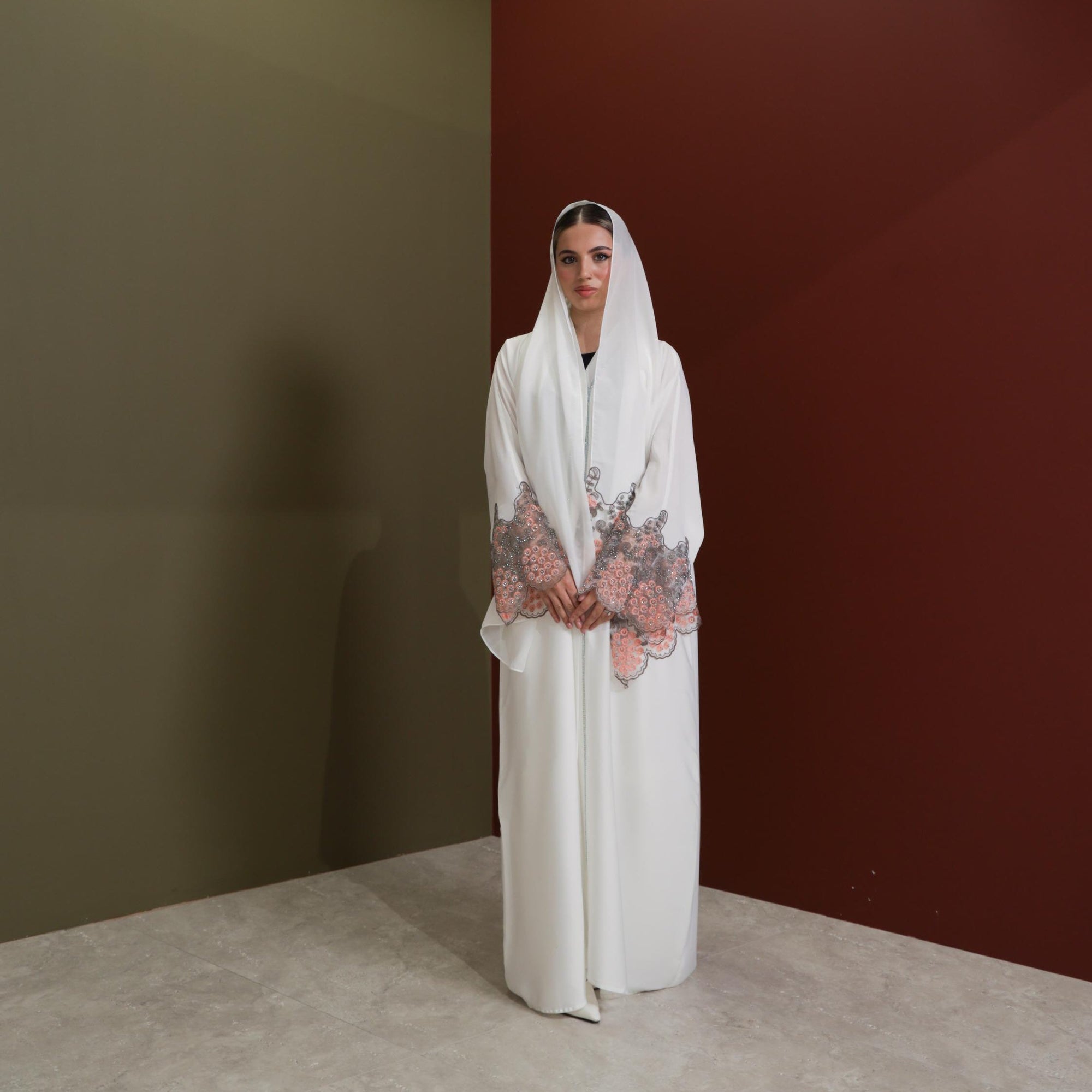 White abaya with silver beadwork and pink and brown embroidery on the sleeves. Made from crepe material with popper buttons, available in sizes 50-60, and includes a matching hijab.