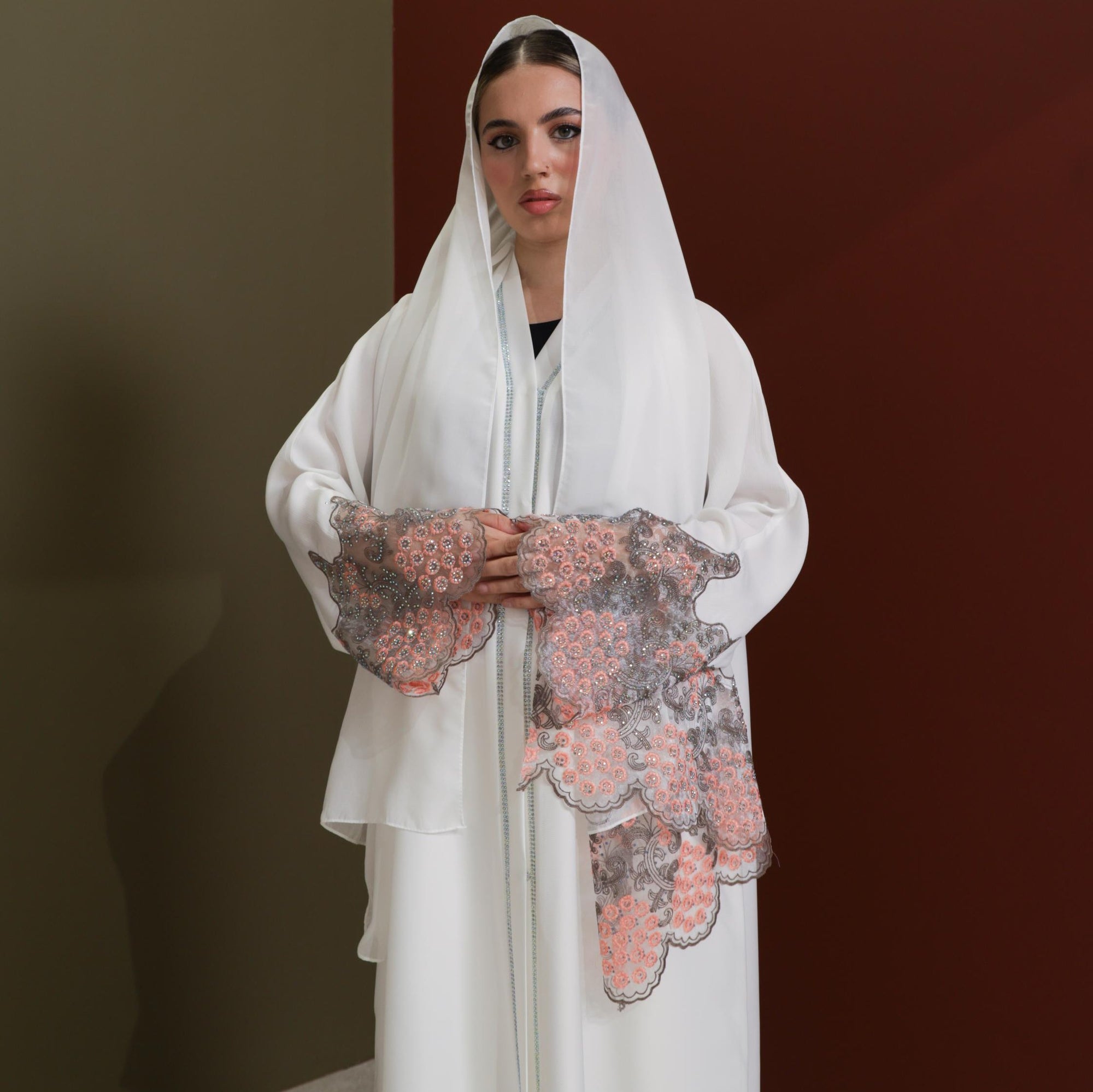 White abaya with silver beadwork and pink and brown embroidery on the sleeves. Made from crepe material with popper buttons, available in sizes 50-60, and includes a matching hijab.