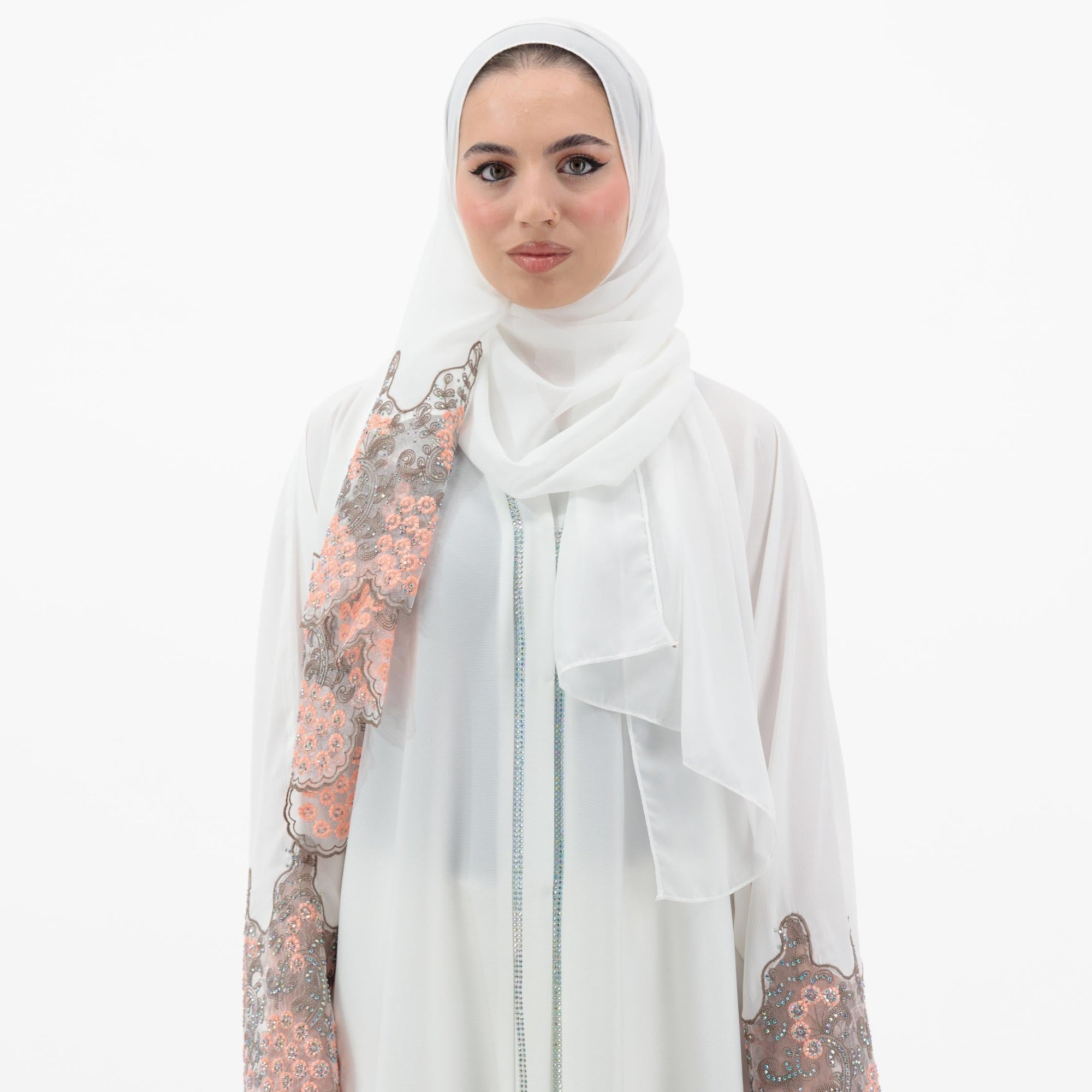 White abaya with silver beadwork and pink and brown embroidery on the sleeves. Made from crepe material with popper buttons, available in sizes 50-60, and includes a matching hijab.