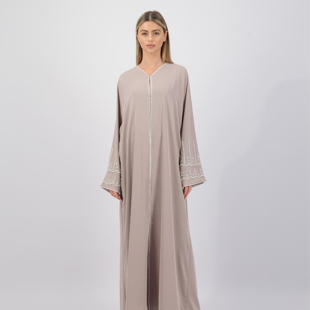 The Sapna Abaya features silver beaded sleeves, popper buttons, and a white line detail. Available in black, beige, brown, and white, made from Zoom fabric in sizes 50-60. Includes a matching hijab.