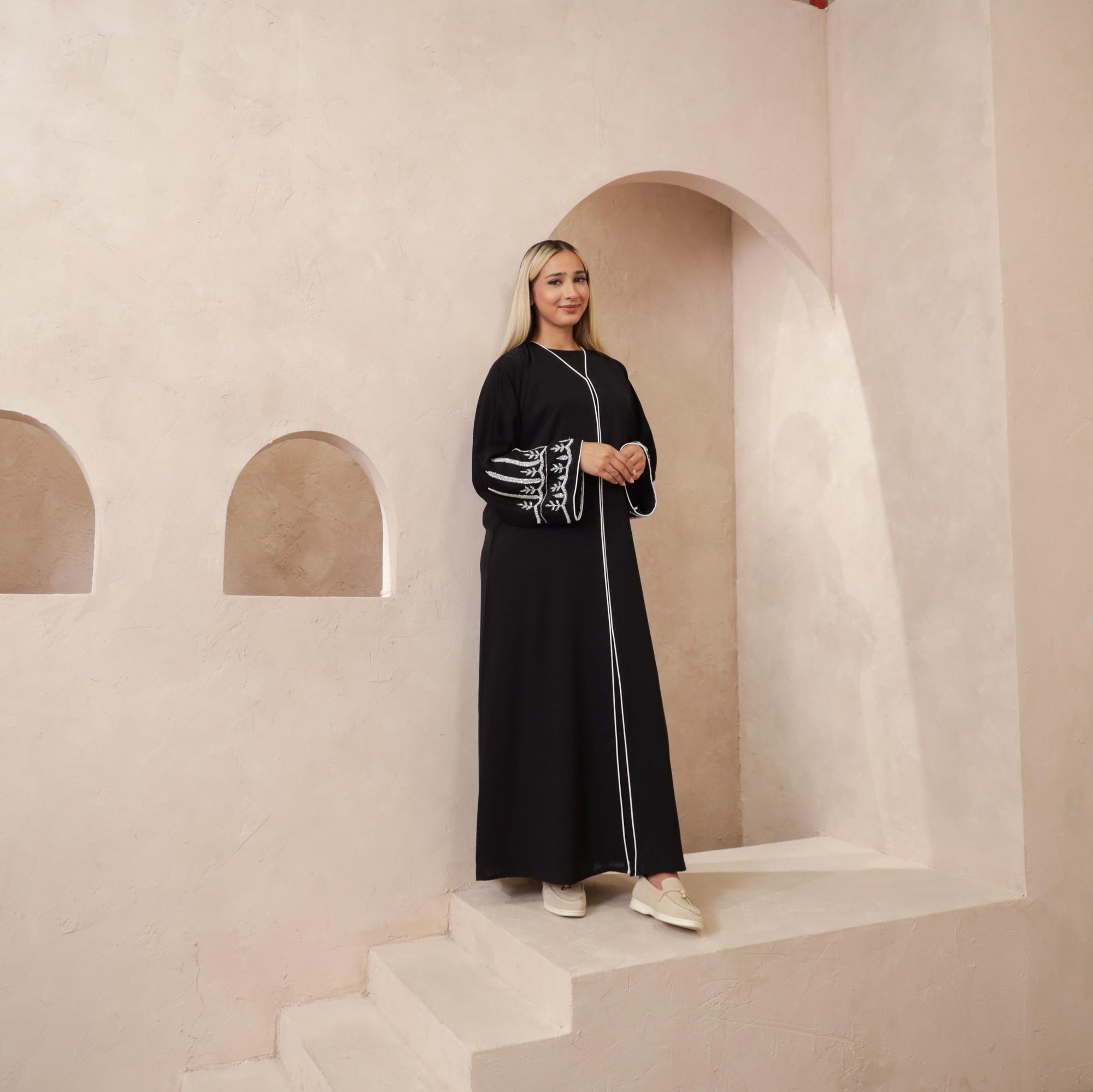 The Sapna Abaya features silver beaded sleeves, popper buttons, and a white line detail. Available in black, beige, brown, and white, made from Zoom fabric in sizes 50-60. Includes a matching hijab.