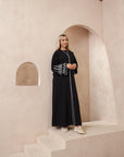 The Sapna Abaya features silver beaded sleeves, popper buttons, and a white line detail. Available in black, beige, brown, and white, made from Zoom fabric in sizes 50-60. Includes a matching hijab.