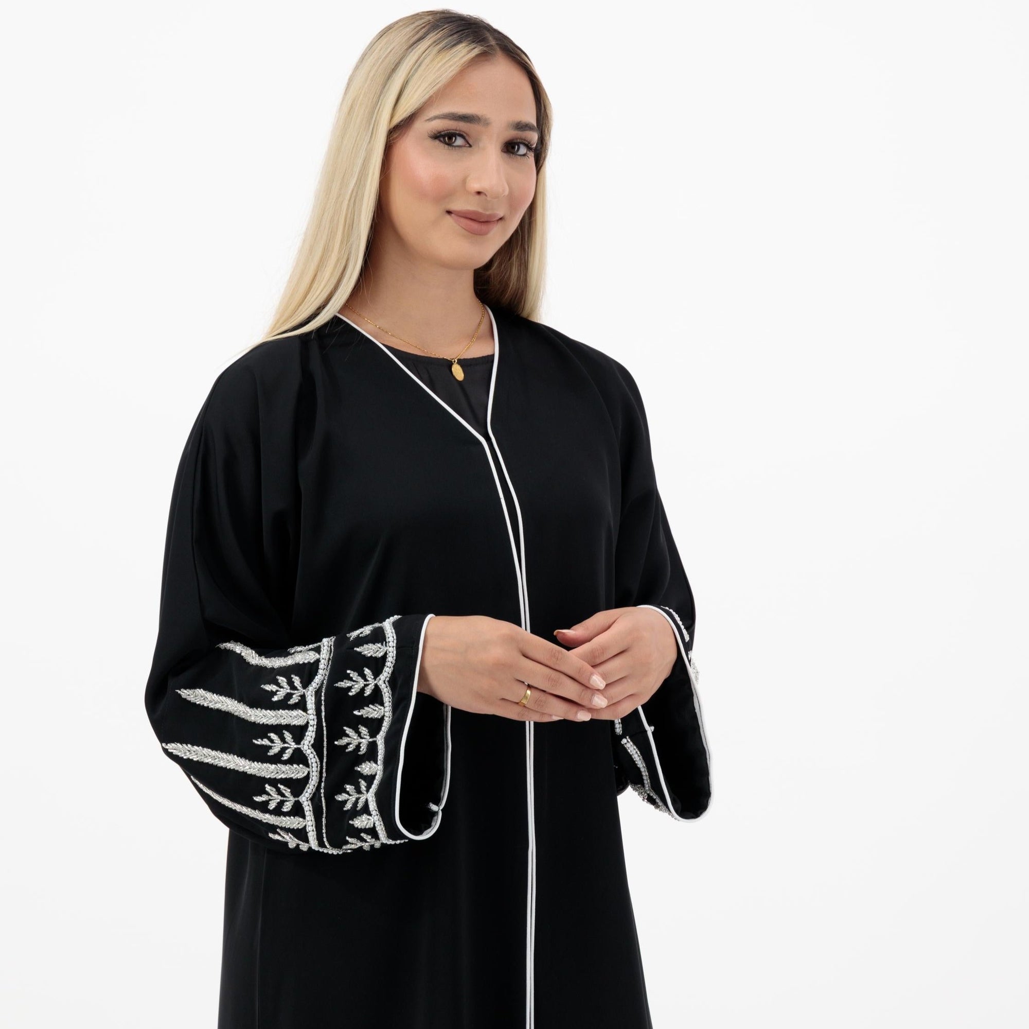 The Sapna Abaya features silver beaded sleeves, popper buttons, and a white line detail. Available in black, beige, brown, and white, made from Zoom fabric in sizes 50-60. Includes a matching hijab.