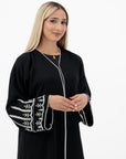 The Sapna Abaya features silver beaded sleeves, popper buttons, and a white line detail. Available in black, beige, brown, and white, made from Zoom fabric in sizes 50-60. Includes a matching hijab.