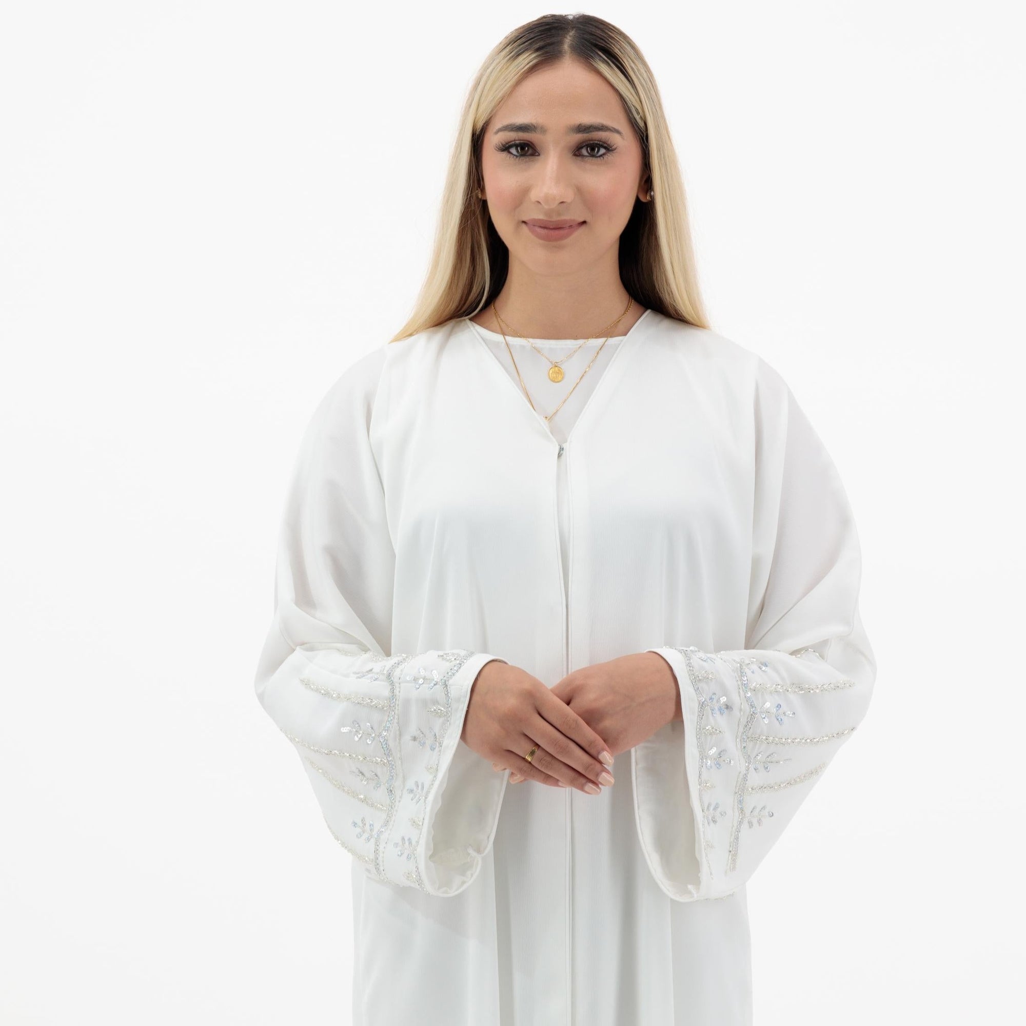 The Sapna Abaya features silver beaded sleeves, popper buttons, and a white line detail. Available in black, beige, brown, and white, made from Zoom fabric in sizes 50-60. Includes a matching hijab.